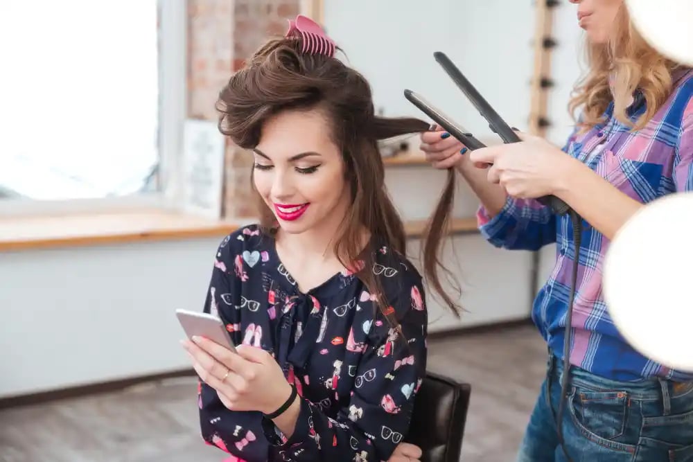 Female hairdresser applying hair straightener for long hair of smiling young woman using smartphone in dressing room_11zon