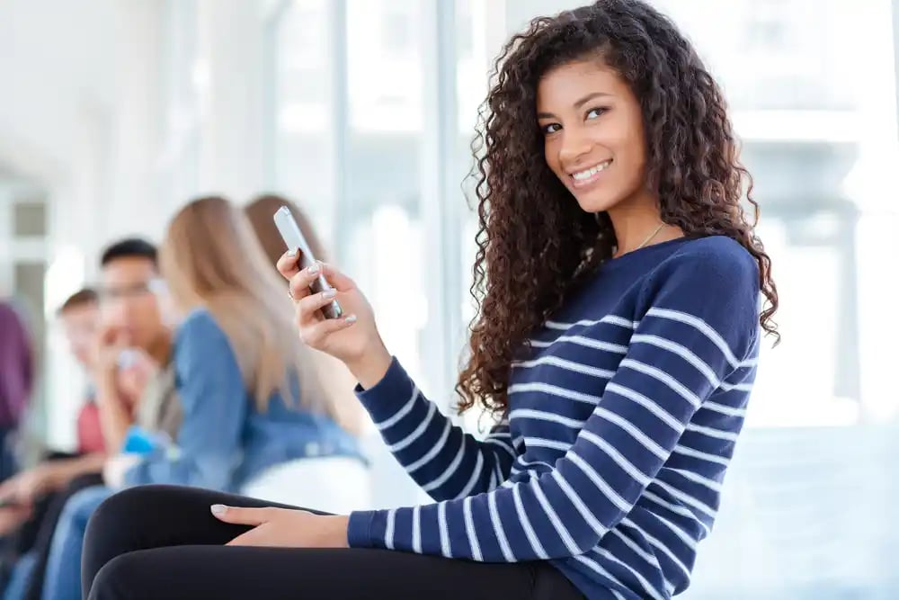 Portrait of a happy afro american female student using smartphone in university hall and looking at camera-1_11zon